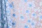 a close-up view of a fabric with a floral pattern, predominantly in shades of blue and white. The fabric appears to be made of a sheer material, with delicate blue flowers and leaves scattered across the surface. The background is a light beige color, providing a neutral backdrop that allows the blue and white floral pattern to stand out.