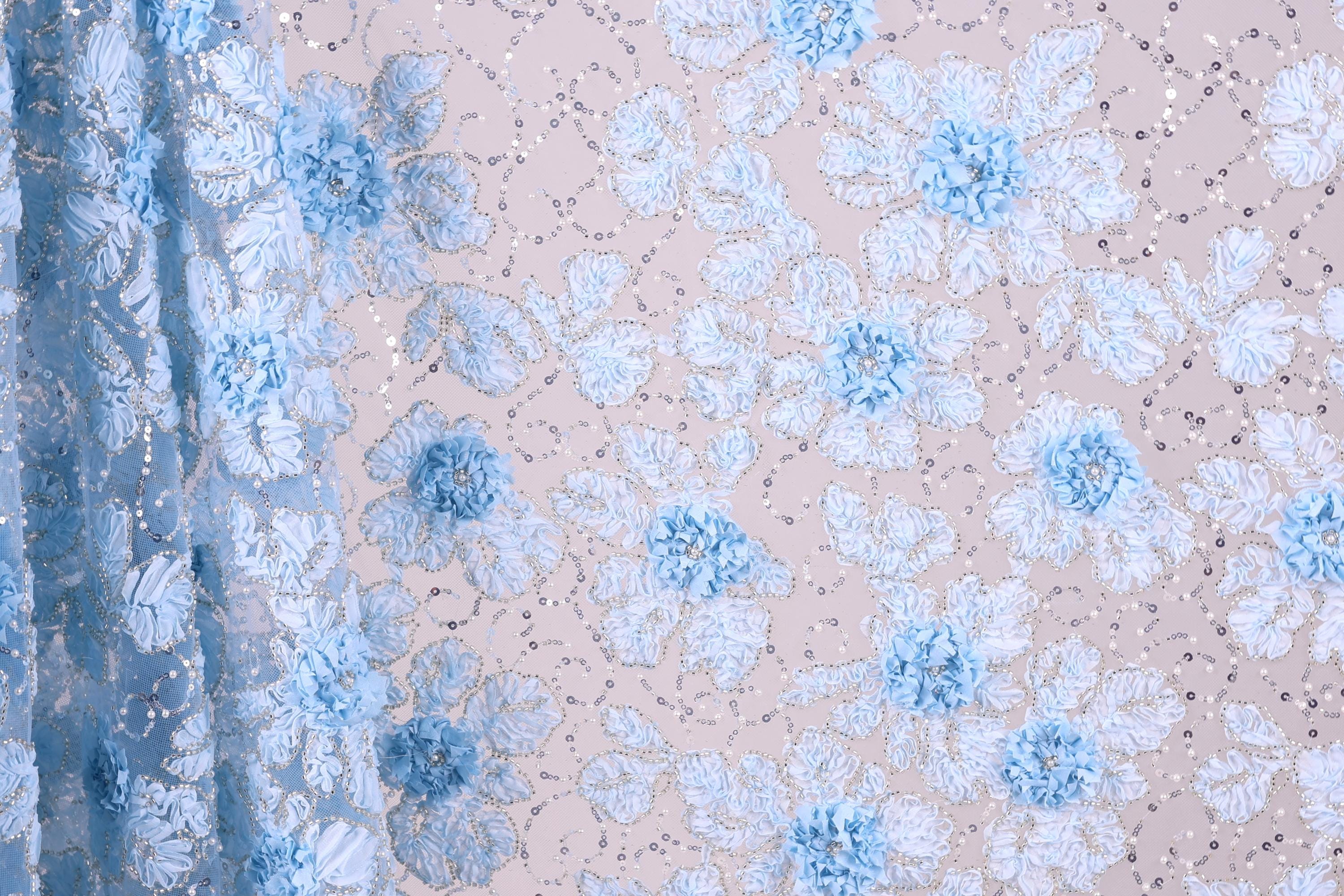 a close-up view of a fabric with a floral pattern, predominantly in shades of blue and white. The fabric appears to be made of a sheer material, with delicate blue flowers and leaves scattered across the surface. The background is a light beige color, providing a neutral backdrop that allows the blue and white floral pattern to stand out.
