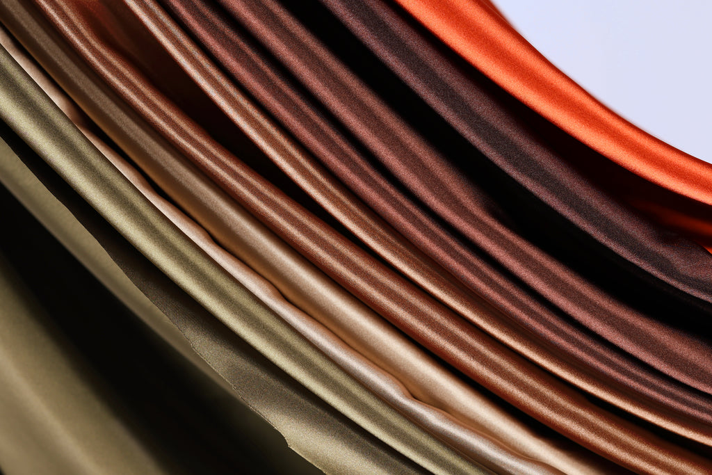 a close-up view of a collection of colorful, shiny fabrics or materials, with a gradient of colors ranging from dark brown to light brown. The fabrics appear to be folded or draped in a way that creates a visually striking and textured effect.