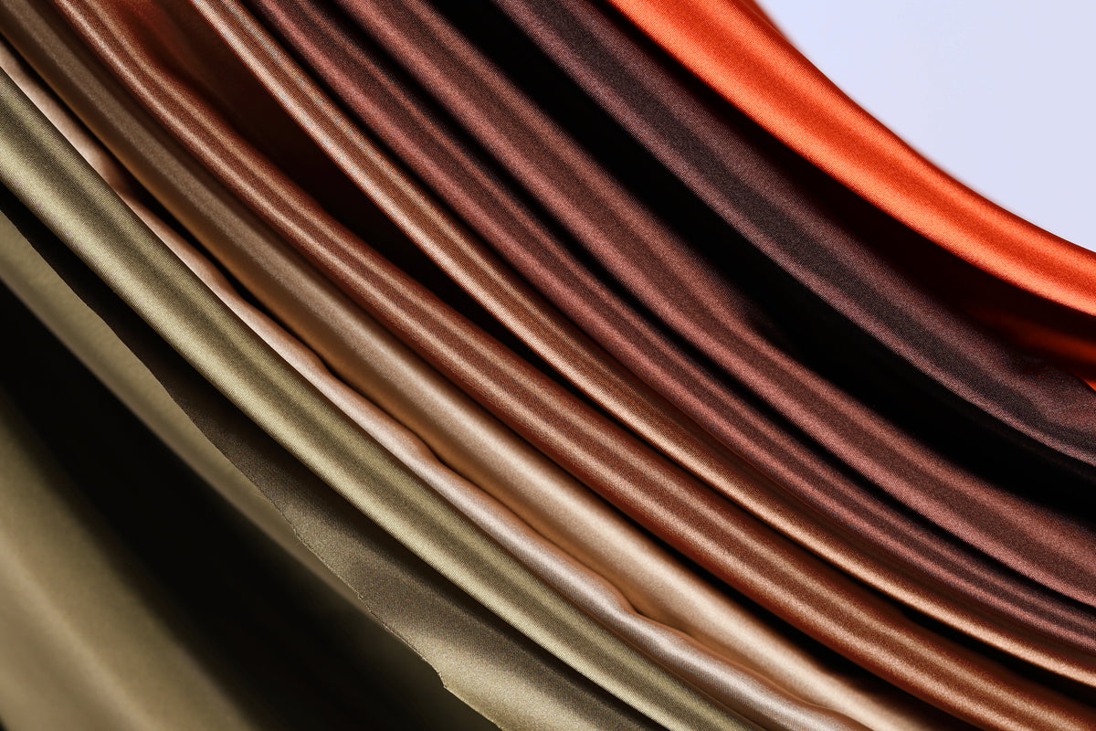a close-up view of a collection of colorful, shiny fabrics or materials, with a gradient of colors ranging from dark brown to light brown. The fabrics appear to be folded or draped in a way that creates a visually striking and textured effect.