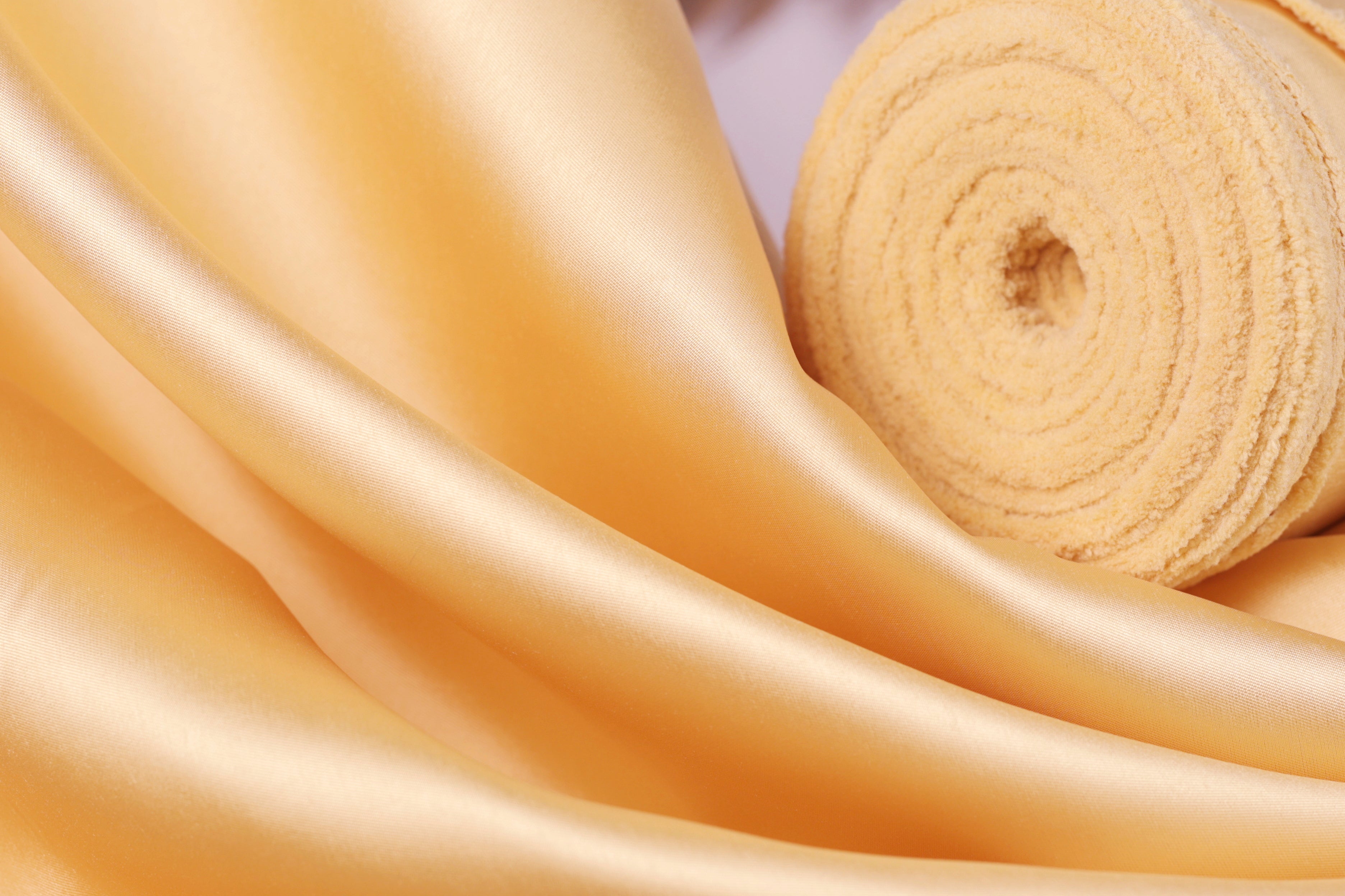 a close-up view of a soft, shiny fabric with a golden hue, possibly silk or satin. The fabric appears to be folded or rolled up, creating a visually appealing texture.