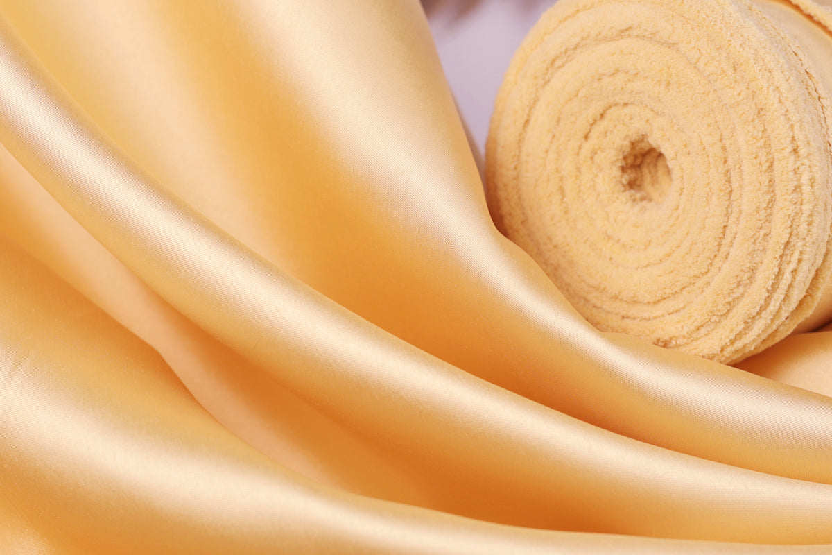 a close-up view of a soft, shiny fabric with a golden hue, possibly silk or satin. The fabric appears to be folded or rolled up, creating a visually appealing texture.