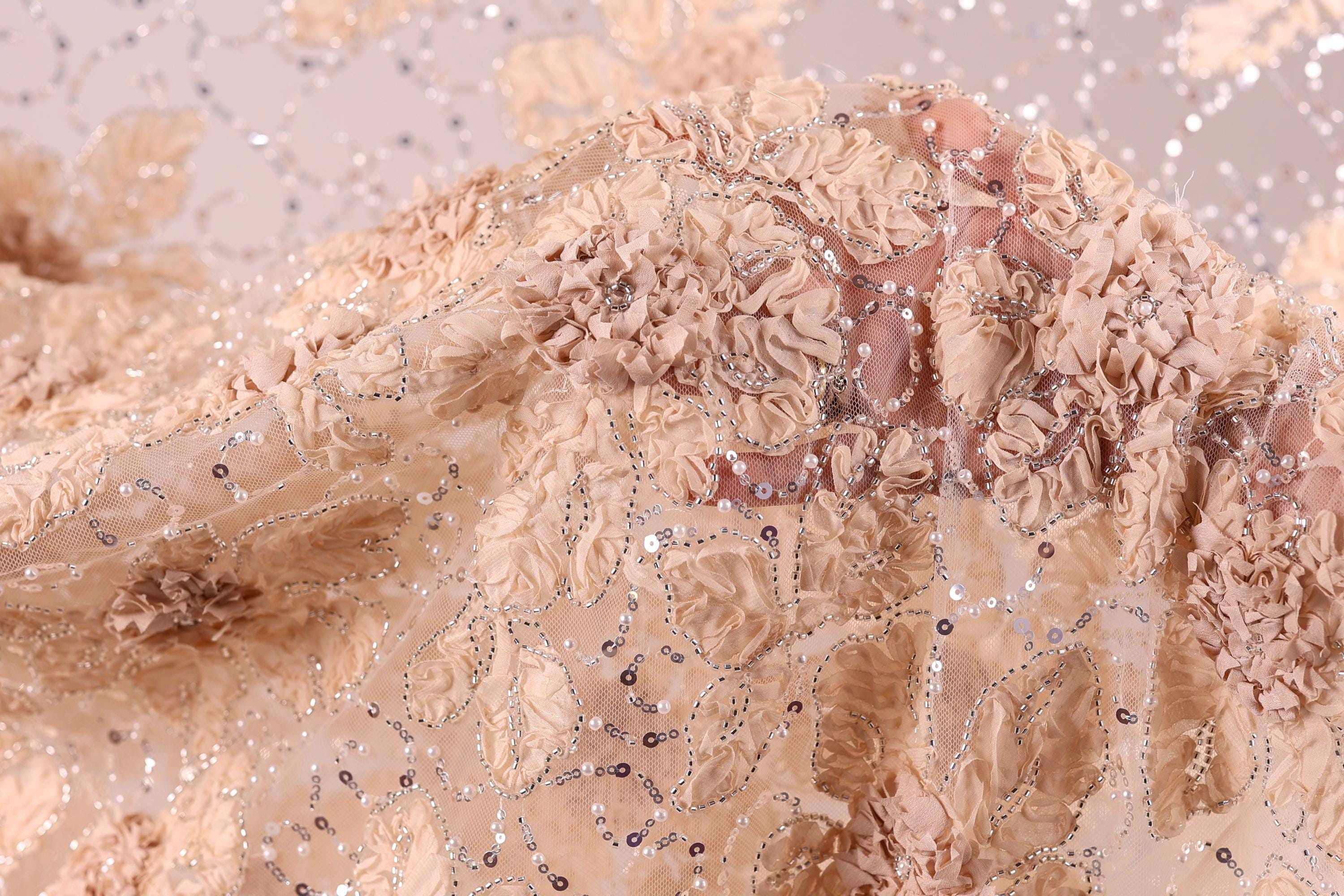 a close-up view of a delicate, intricately embroidered fabric with a floral pattern. The fabric appears to be made of a light-colored material, possibly lace or chiffon, and is adorned with numerous small beads or sequins that add a touch of sparkle and detail to the design. The image does not contain any text or other discernible objects, and the focus is solely on the fabric's intricate details and textures.
