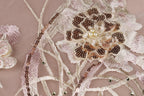 The image is a close-up of a delicate, intricately embroidered fabric with a floral design. The fabric features a central flower-like motif surrounded by smaller, circular embellishments, such as sequins and pearls. The overall composition is visually appealing and showcases the craftsmanship of the embroidery.