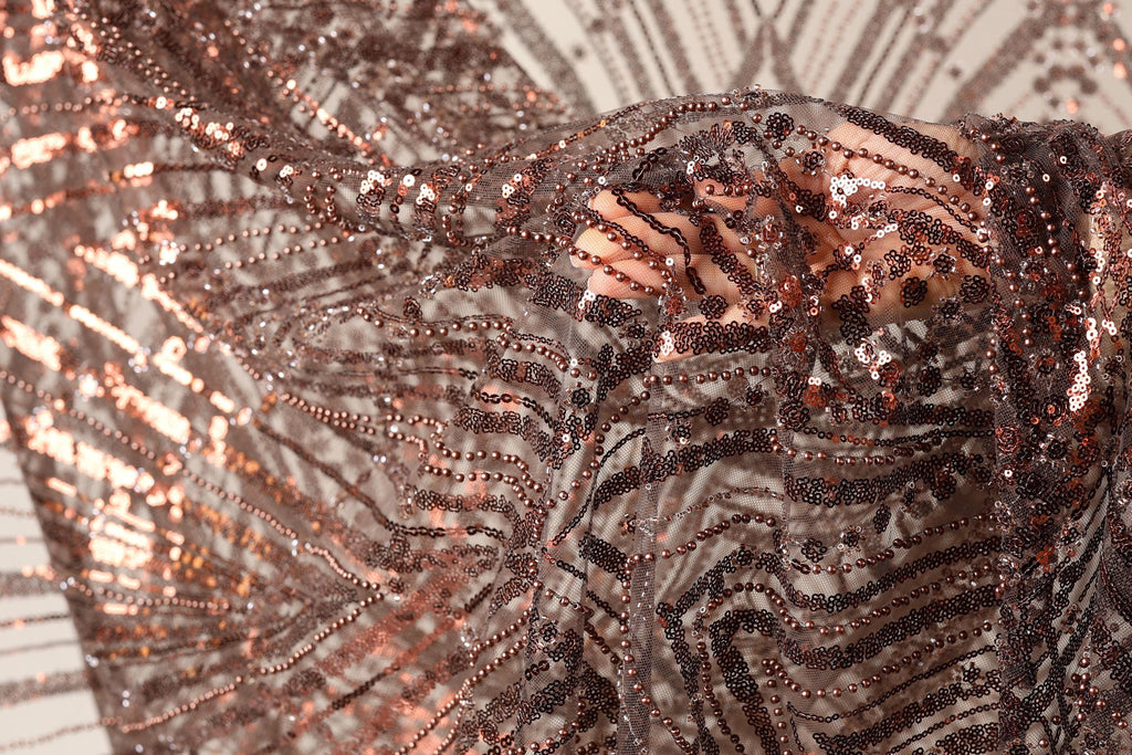 The image is a close-up of a piece of fabric with a metallic, sequined texture. The fabric appears to be made of a shiny material, possibly metallic or sequin-like, and has a pattern of interlocking lines and dots. The fabric is being held up by a hand, creating a sense of movement and texture. The background is blurred, drawing focus to the fabric and its details.
