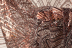 The image is a close-up of a piece of fabric with a metallic, sequined texture. The fabric appears to be made of a shiny material, possibly metallic or sequin-like, and has a pattern of interlocking lines and dots. The fabric is being held up by a hand, creating a sense of movement and texture. The background is blurred, drawing focus to the fabric and its details.
