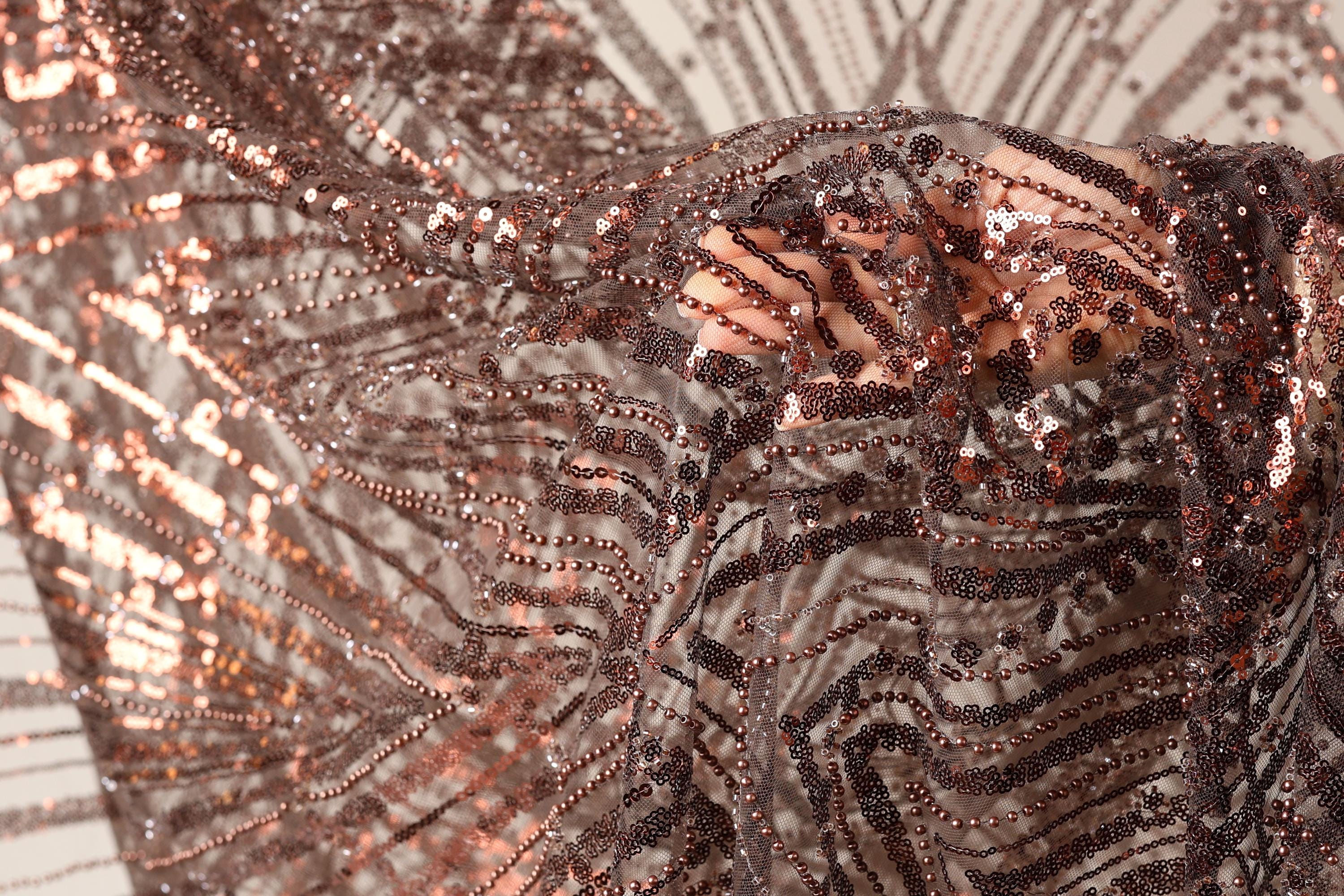 The image is a close-up of a piece of fabric with a metallic, sequined texture. The fabric appears to be made of a shiny material, possibly metallic or sequin-like, and has a pattern of interlocking lines and dots. The fabric is being held up by a hand, creating a sense of movement and texture. The background is blurred, drawing focus to the fabric and its details.