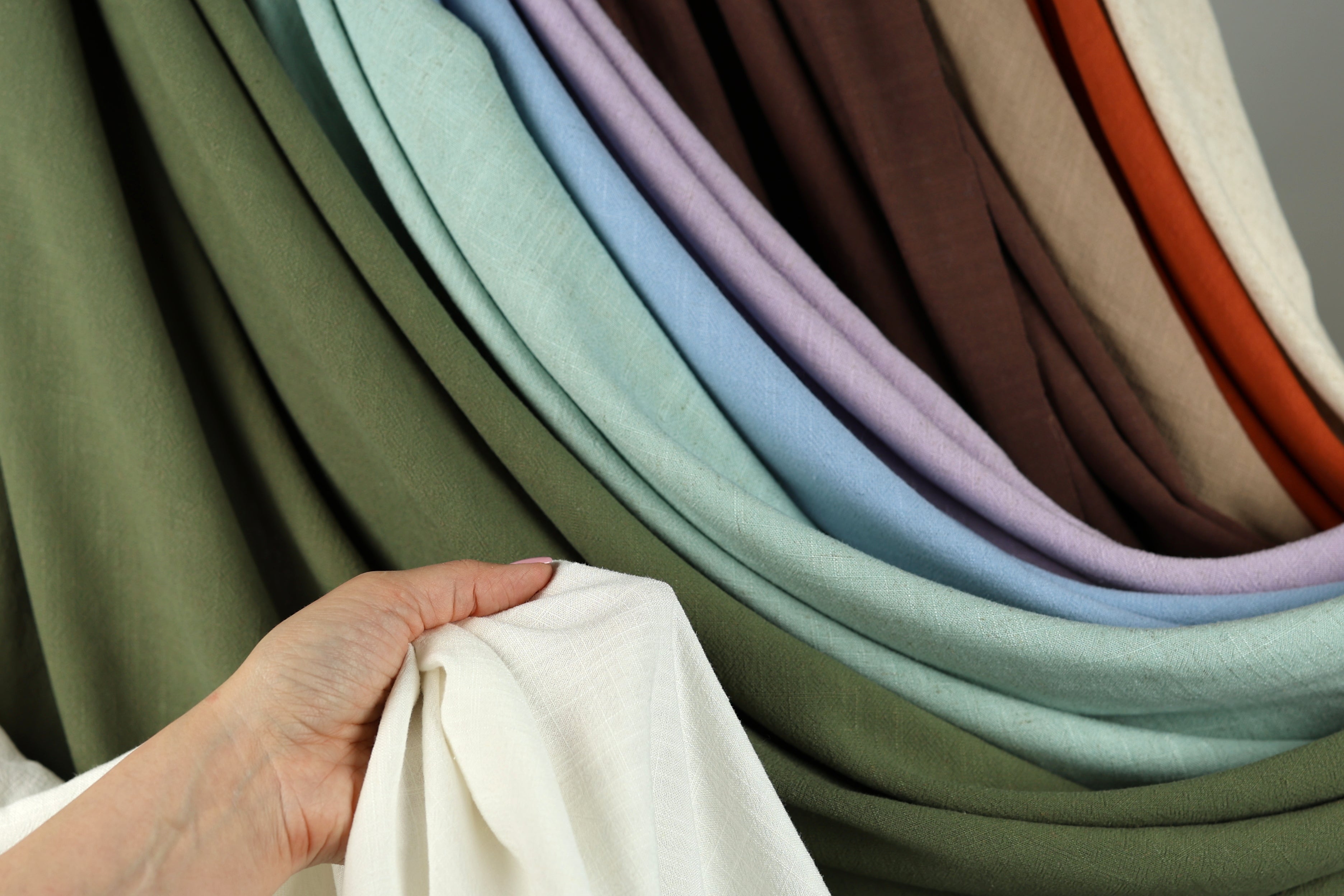 A hand is holding a white fabric in front of a colorful array of fabric swatches, showcasing various shades and patterns.