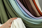 A hand is holding a white fabric in front of a colorful array of fabric swatches, showcasing various shades and patterns.