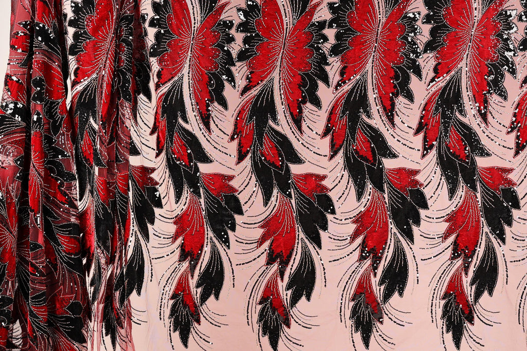 a close-up view of a fabric with a striking pattern of red and black feathers or leaves. The pattern is intricate and detailed, with the feathers or leaves appearing to be made of sequins or beads, adding a touch of sparkle to the design. The fabric is predominantly pink, providing a contrasting background for the bold red and black elements. The image does not contain any text or other discernible objects, and the focus is solely on the fabric and its captivating design.