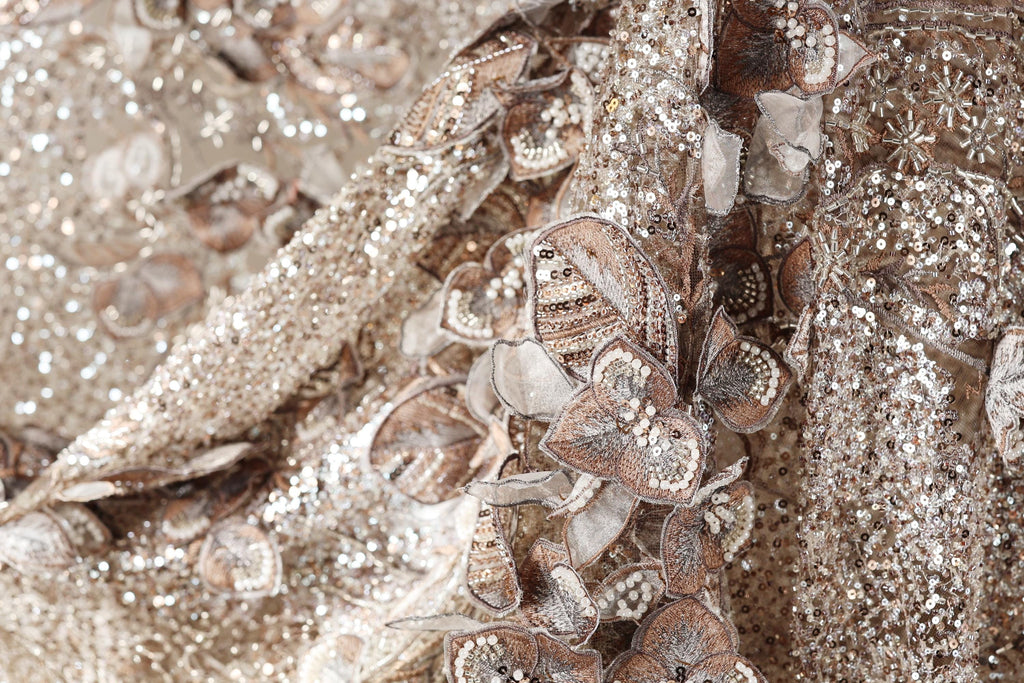 The image is a close-up of a fabric with a gold and brown color scheme, featuring intricate embroidered designs and sequins.