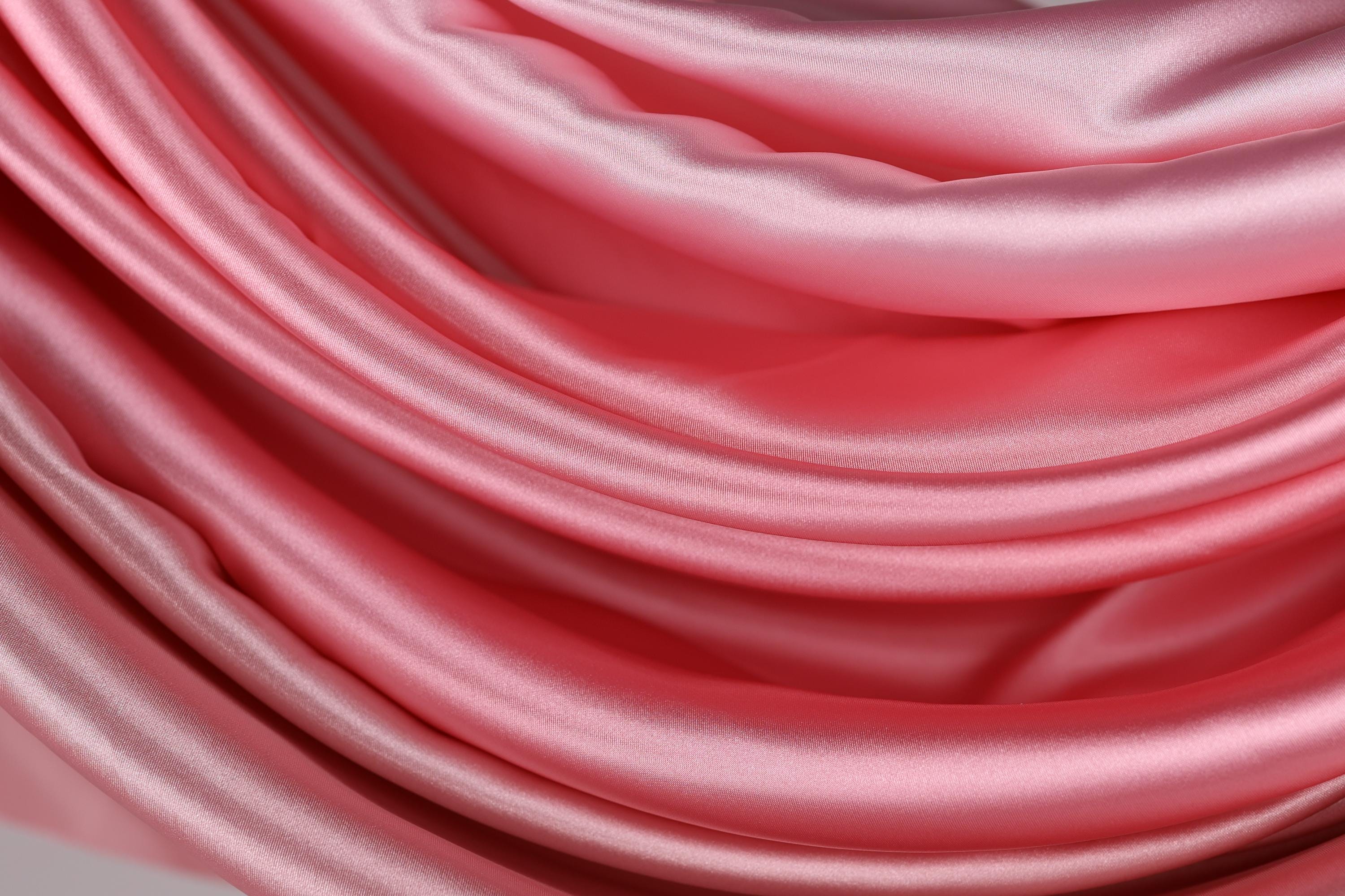 The image is a close-up of a soft, shiny pink satin fabric, with its folds and creases creating a textured and flowing appearance.