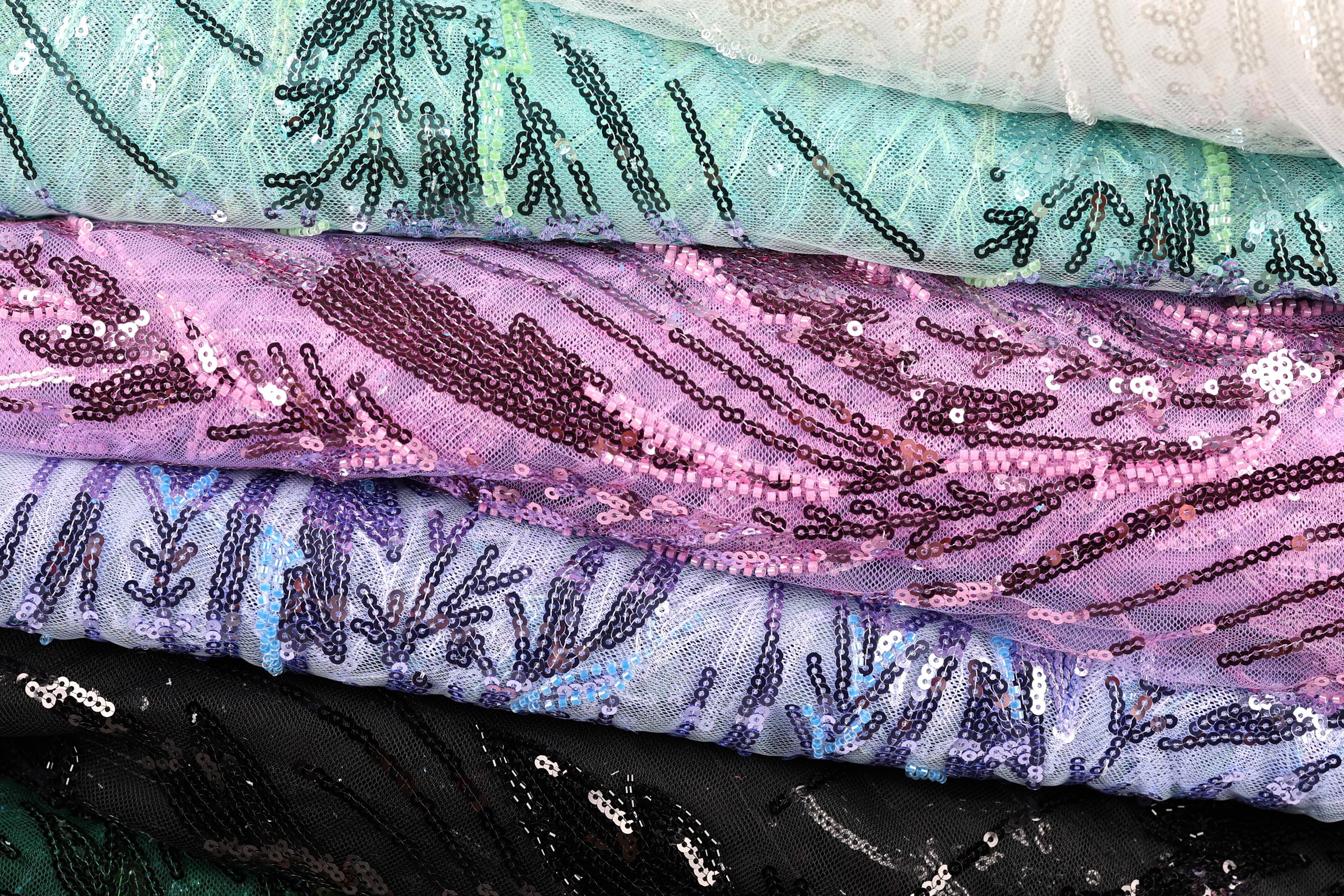 a close-up view of several colorful fabric pieces, including a purple and pink fabric with sequins, a white fabric with sequins, and a black fabric with sequins. The fabrics appear to be folded or stacked together, creating a visually appealing and textured composition.