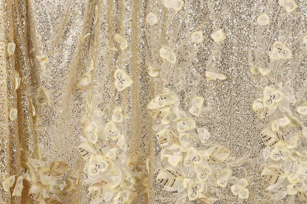 The image is a close-up of a fabric with a gold and white floral pattern. The fabric appears to be made of a shiny, sequined material, giving it a luxurious and elegant appearance.