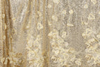 The image is a close-up of a fabric with a gold and white floral pattern. The fabric appears to be made of a shiny, sequined material, giving it a luxurious and elegant appearance.