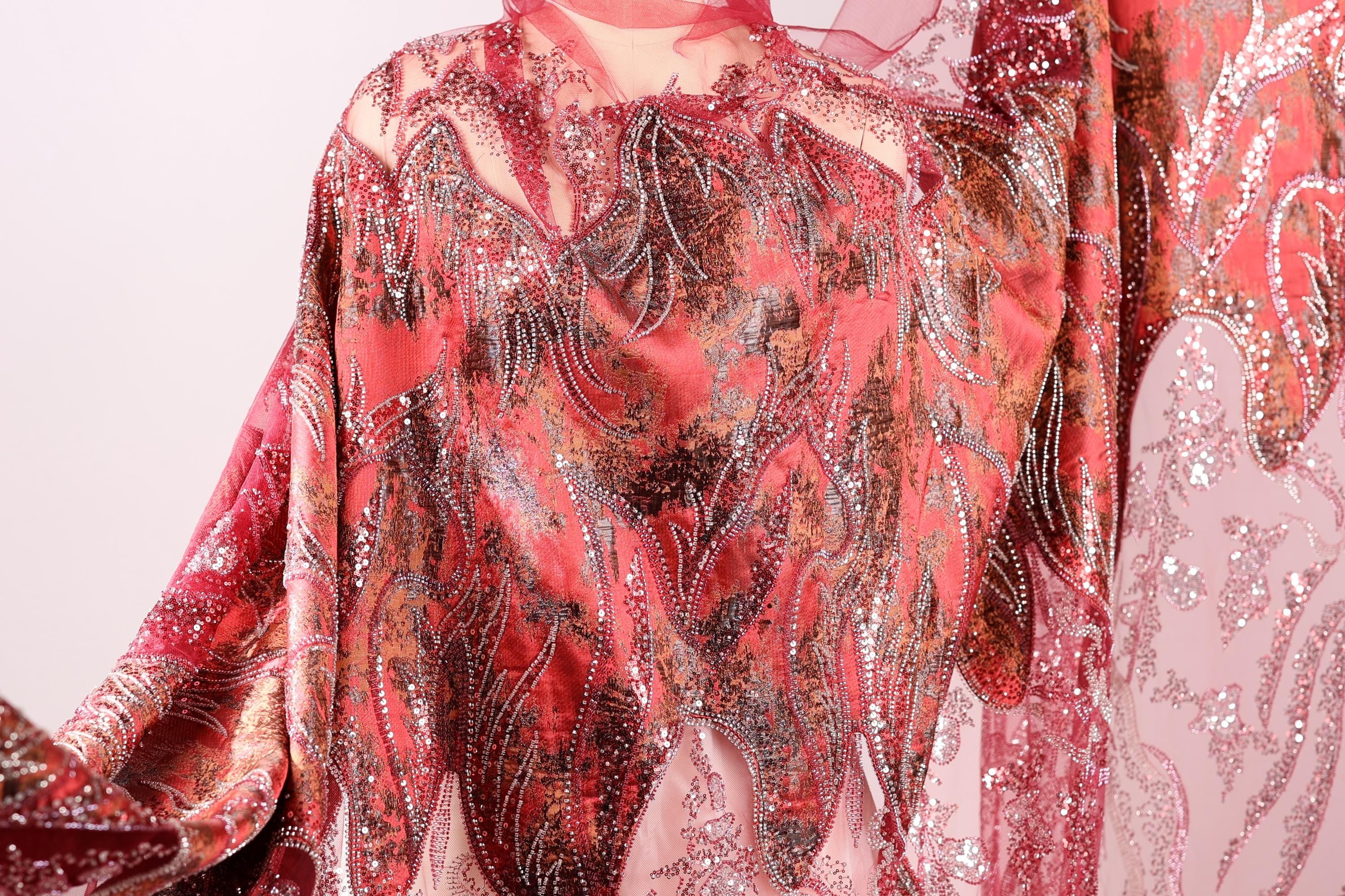 a close-up view of a vibrant, patterned fabric with a mix of red, pink, and orange colors. The fabric appears to be made of a shiny, sequined material, giving it a luxurious and eye-catching appearance.