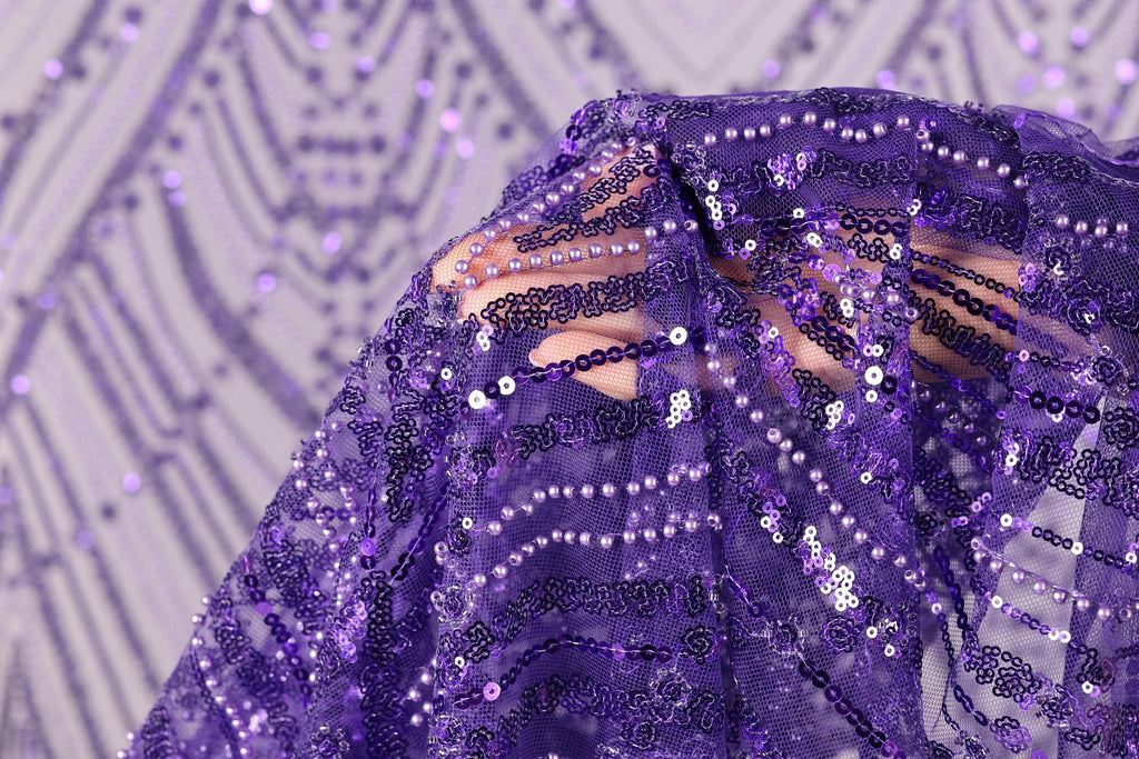 A close-up view of a purple sequin fabric, with a blurred background that appears to be a patterned fabric or a wall.