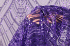 A close-up view of a purple sequin fabric, with a blurred background that appears to be a patterned fabric or a wall.
