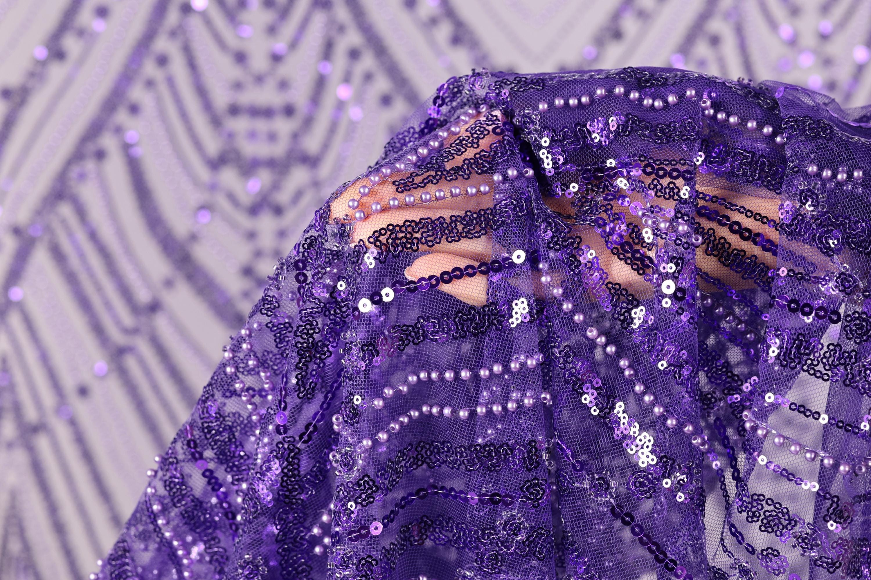 A close-up view of a purple sequin fabric, with a blurred background that appears to be a patterned fabric or a wall.