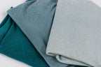 a close-up view of three fabric swatches, each with a different color and texture. The swatches are arranged in a way that they overlap each other, creating a layered effect. The colors of the swatches are teal, blue, and gray, with the teal and blue colors being more prominent. The gray color is also visible in the image.
