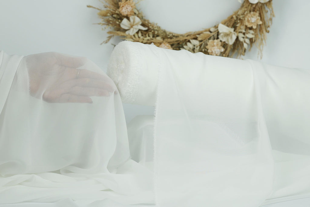 a hand holding a piece of white fabric, with a wreath of flowers in the background.