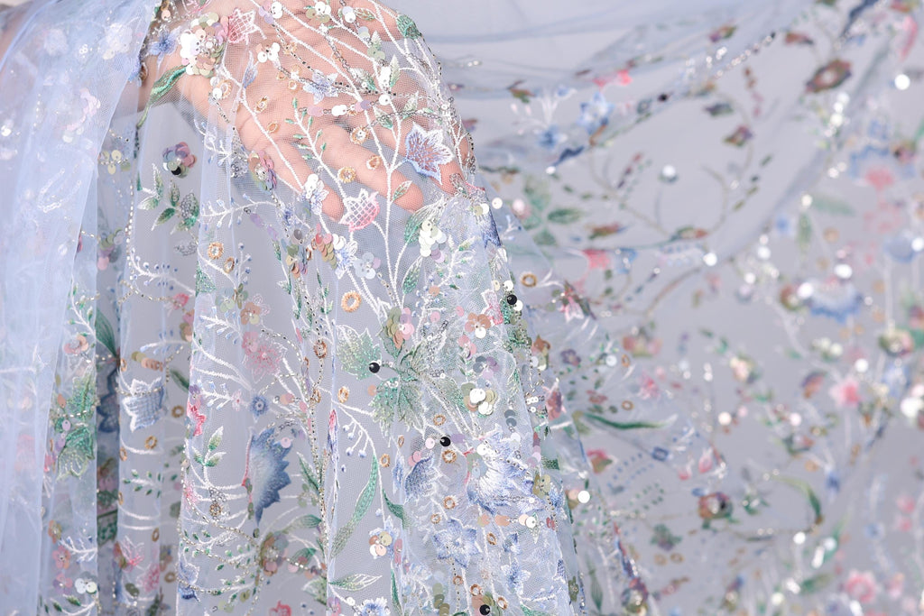 a close-up view of a delicate, intricately embroidered fabric with a floral pattern. The fabric appears to be made of sheer material, giving it a soft and delicate appearance. The embroidery features a variety of flowers and leaves, creating a visually appealing and detailed design. The fabric is spread out, allowing for a clear view of its intricate details and patterns.