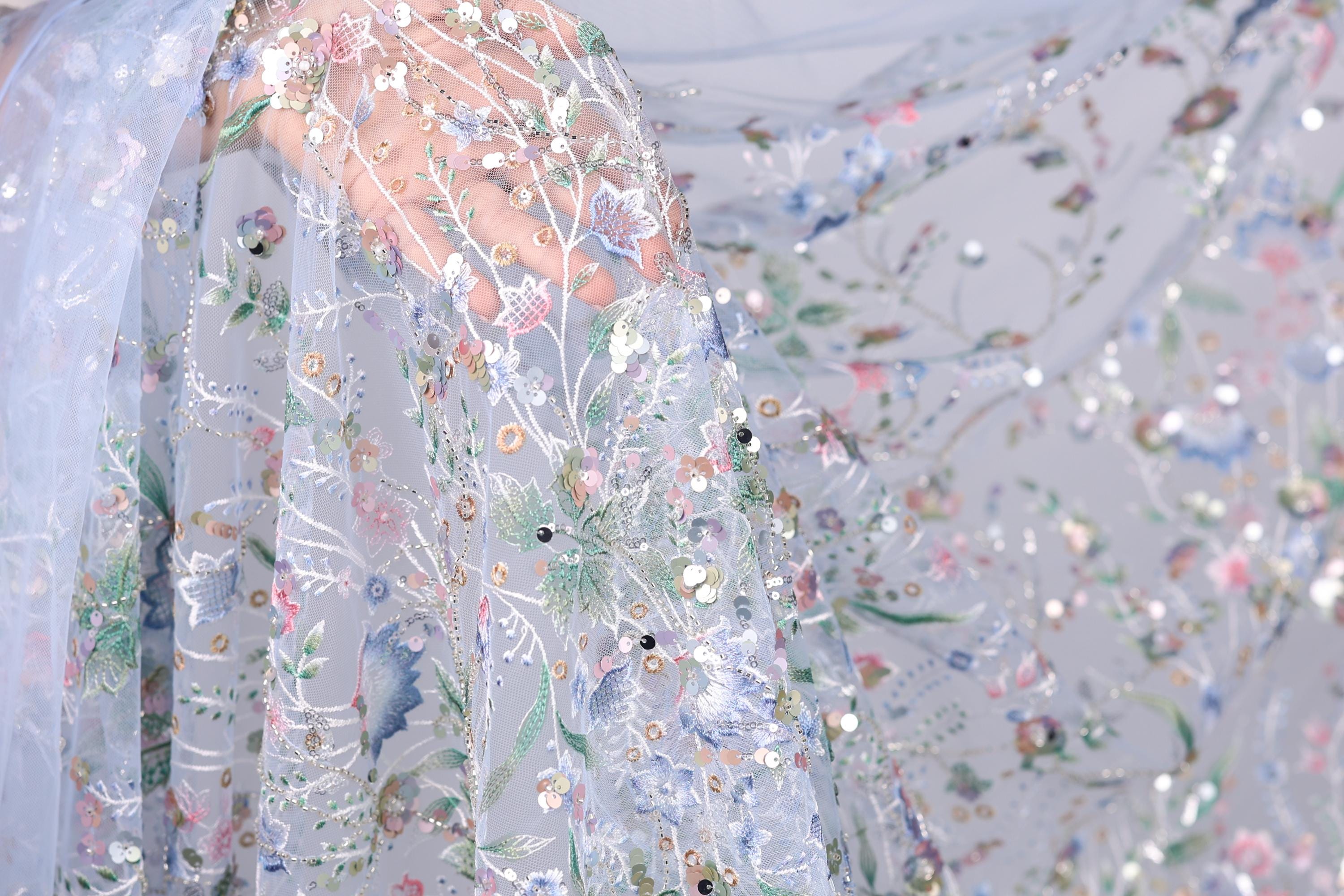 a close-up view of a delicate, intricately embroidered fabric with a floral pattern. The fabric appears to be made of sheer material, giving it a soft and delicate appearance. The embroidery features a variety of flowers and leaves, creating a visually appealing and detailed design. The fabric is spread out, allowing for a clear view of its intricate details and patterns.