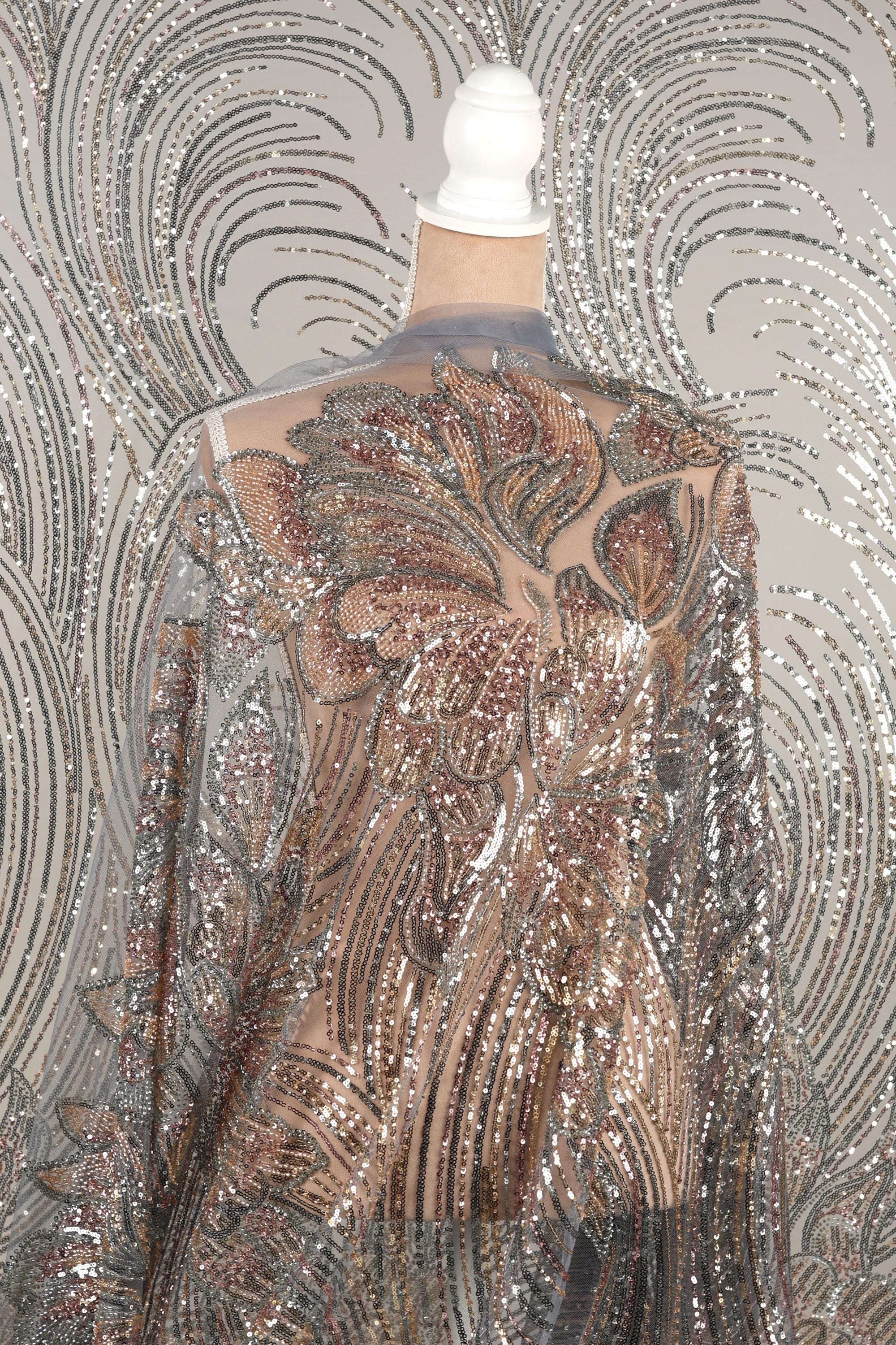 a mannequin wearing a shimmering, sequined dress with a floral pattern. The dress has a sheer back and long sleeves, giving it a delicate and elegant appearance.