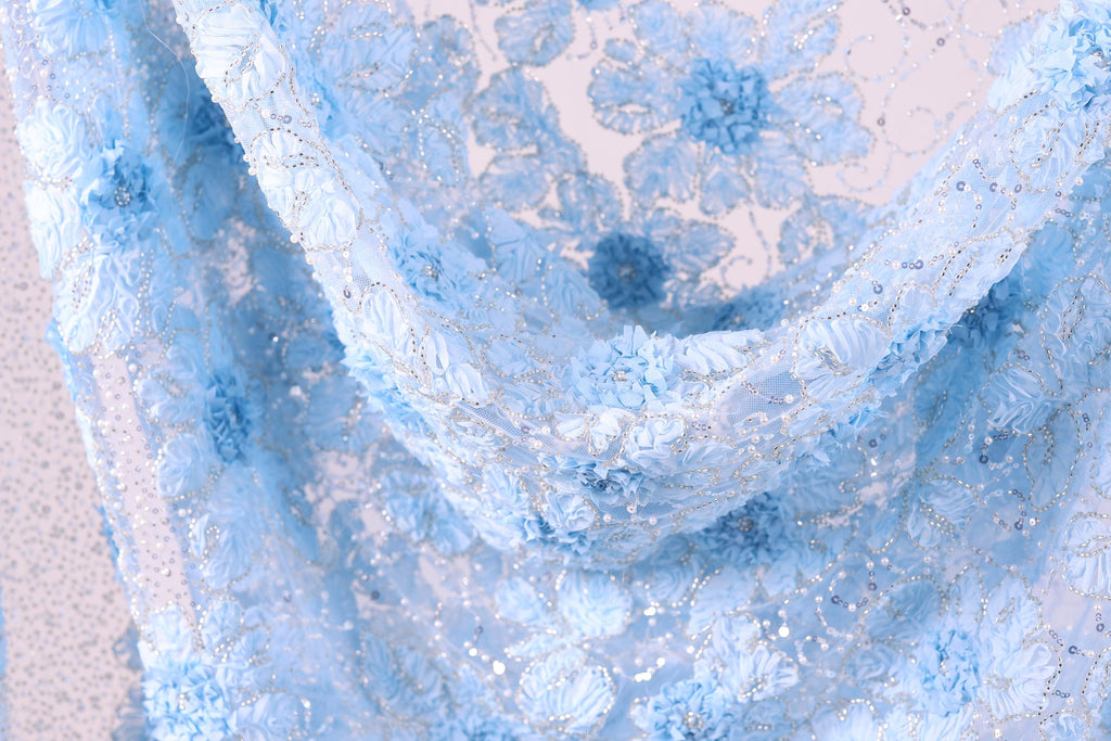 The image is a close-up view of a delicate, blue fabric with a floral pattern. The fabric appears to be made of lace or a similar delicate material, with intricate designs and patterns that create a visually appealing and textured appearance.