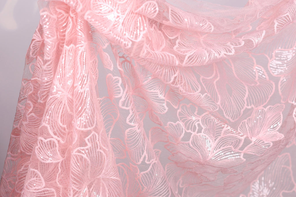 a close-up view of a delicate, pink lace fabric with a floral pattern. The fabric appears to be made of sheer material, giving it a soft and delicate appearance. The lace is intricately designed with a repeating pattern of leaves and flowers, creating a visually appealing and elegant design.