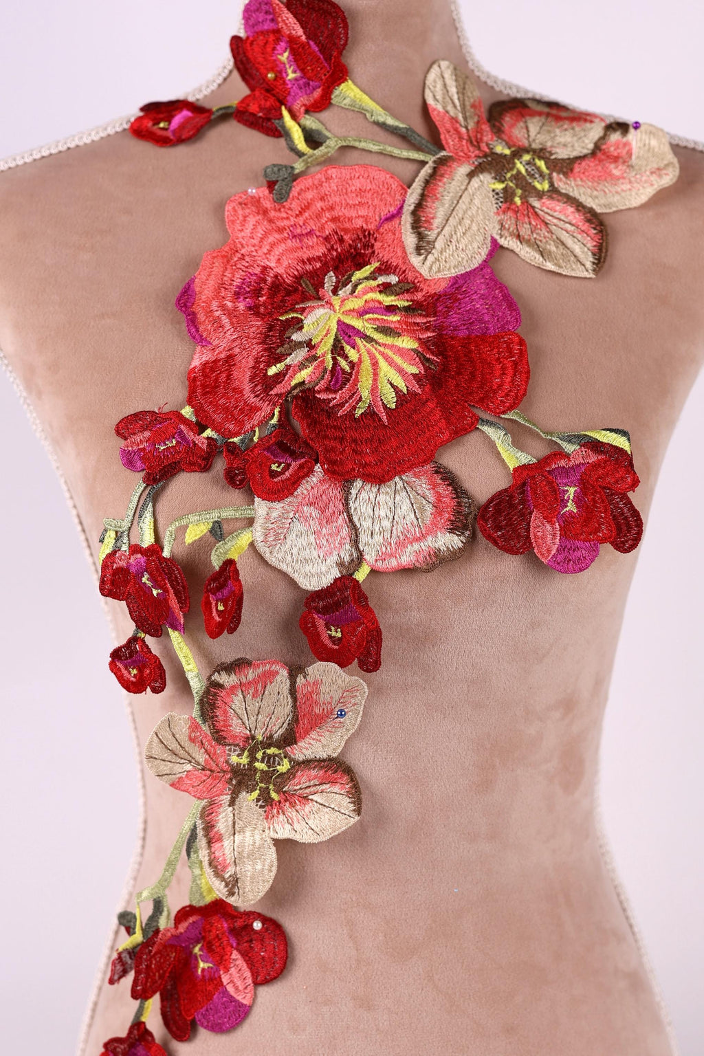 a mannequin with a beige dress adorned with a large, intricately embroidered floral design. The design consists of a variety of red and pink flowers, including a prominent red rose, arranged in a cascading manner. The embroidery appears to be done with a high level of detail and craftsmanship, creating a visually striking and elegant appearance.