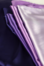a close-up view of a pile of fabric, with a mix of purple and white colors. The fabric appears to be made of a soft, shiny material, possibly silk or satin. The image does not contain any discernible text or specific details about the objects or their arrangement.