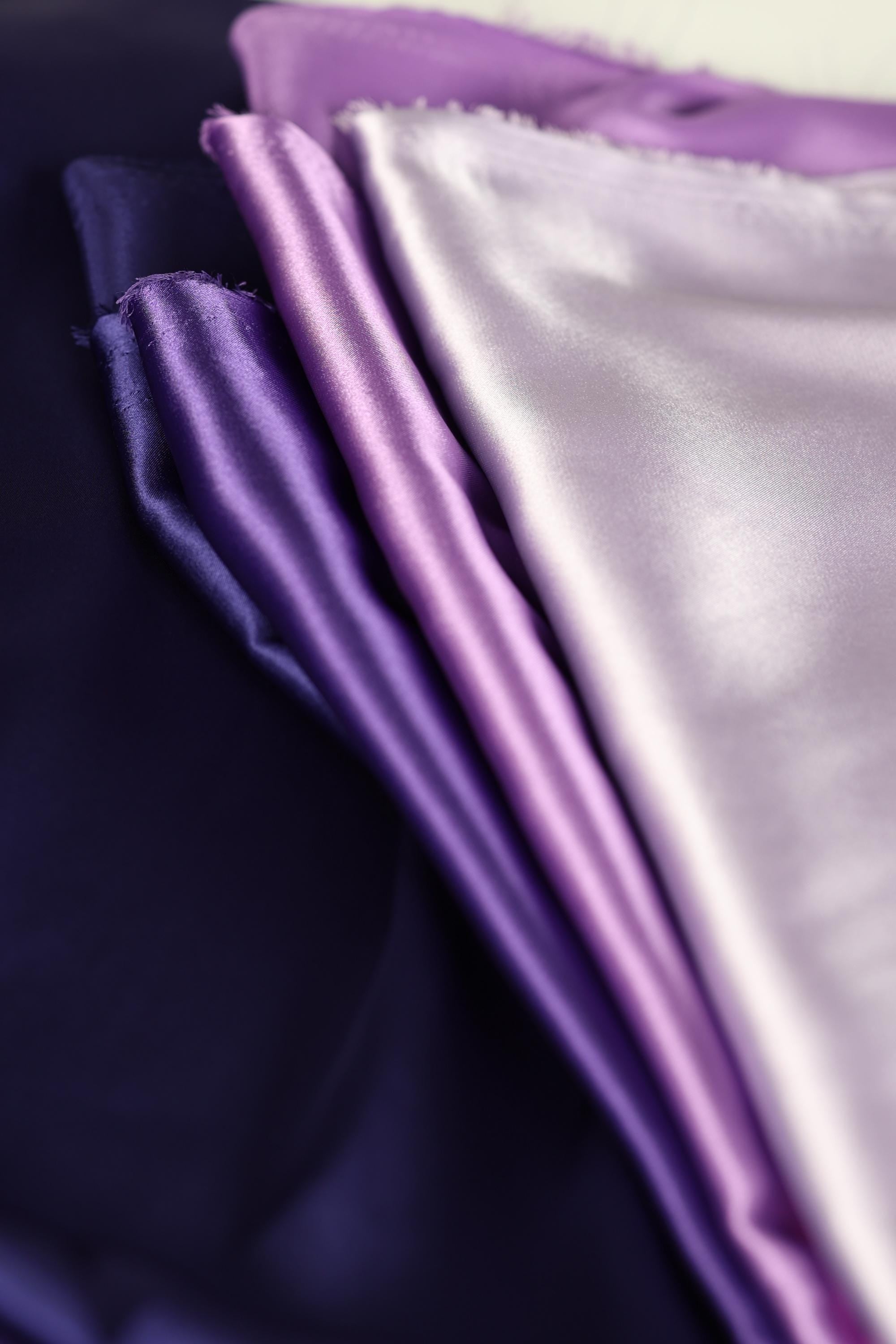 a close-up view of a pile of fabric, with a mix of purple and white colors. The fabric appears to be made of a soft, shiny material, possibly silk or satin. The image does not contain any discernible text or specific details about the objects or their arrangement.