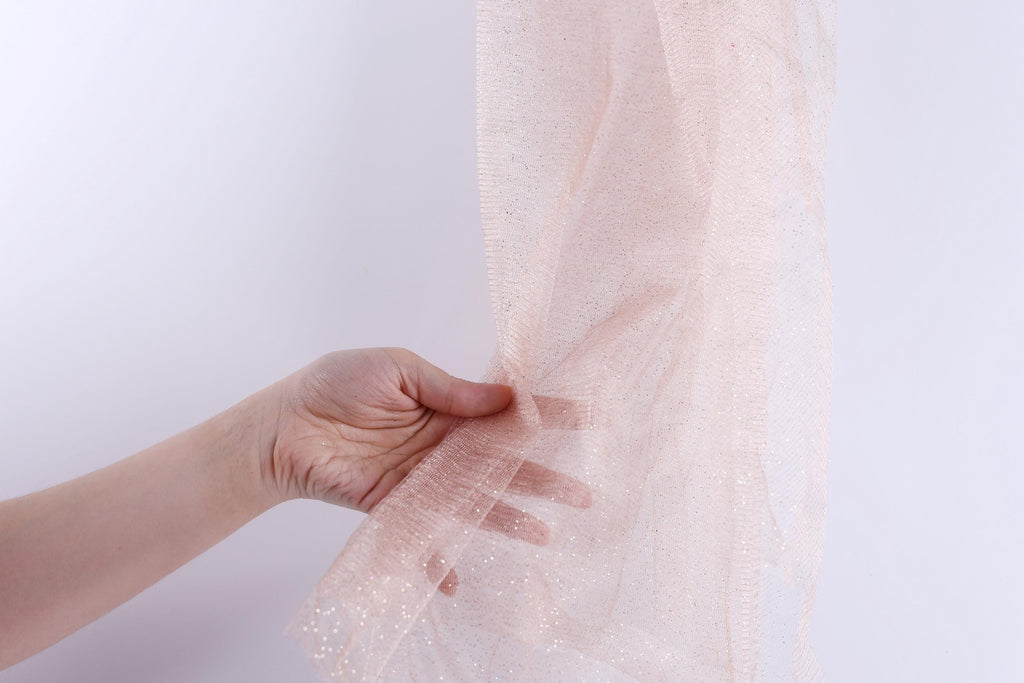 A hand is holding a piece of pink, sparkly fabric against a white background.