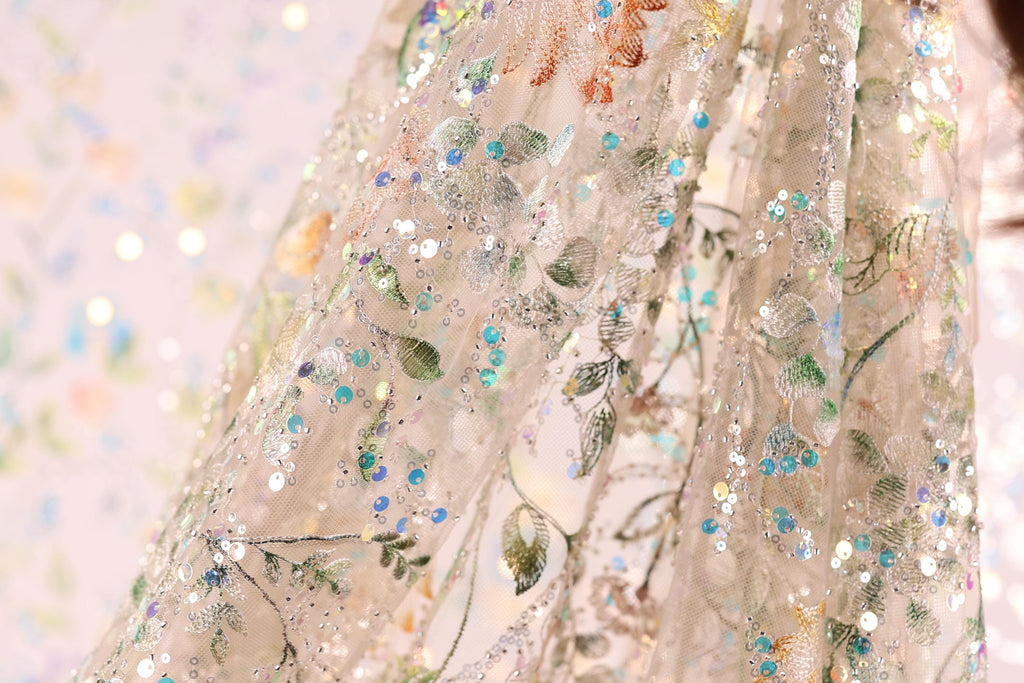a close-up view of a delicate, intricately embroidered fabric with a floral pattern. The fabric appears to be made of sheer material, giving it a delicate and ethereal appearance. The embroidery features a variety of colors and patterns, including green leaves, blue flowers, and white and silver sequins. The fabric is displayed against a blurred background, which emphasizes its intricate details and vibrant colors.