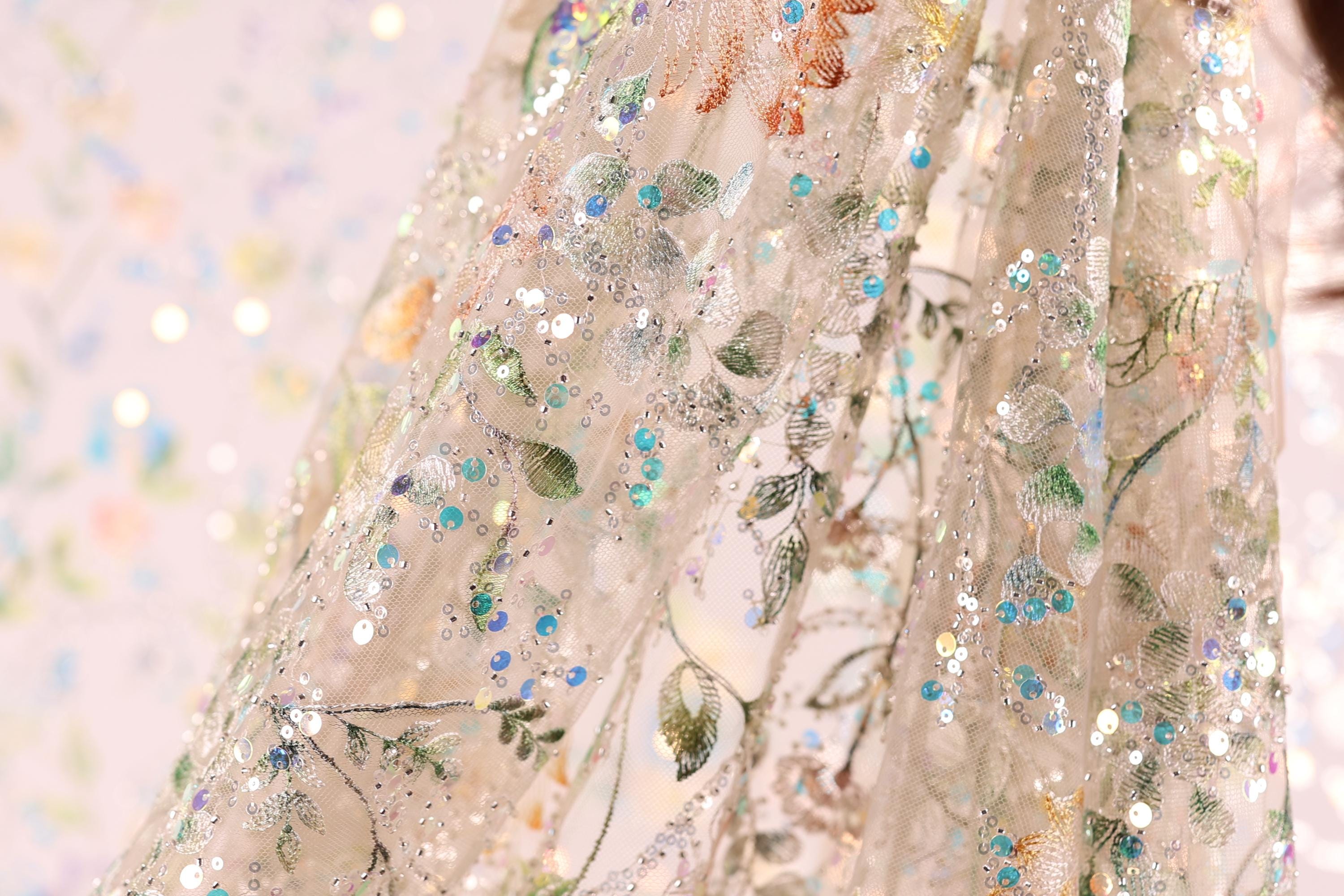 a close-up view of a delicate, intricately embroidered fabric with a floral pattern. The fabric appears to be made of sheer material, giving it a delicate and ethereal appearance. The embroidery features a variety of colors and patterns, including green leaves, blue flowers, and white and silver sequins. The fabric is displayed against a blurred background, which emphasizes its intricate details and vibrant colors.