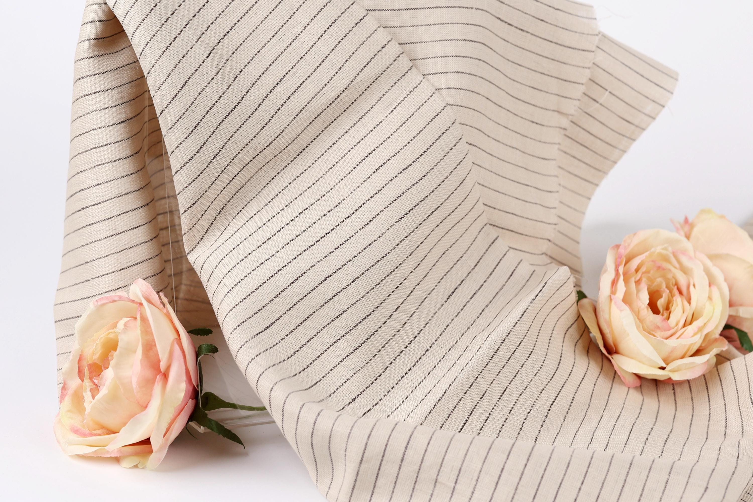 A beige fabric with black stripes is draped over a white background, with two delicate pink roses resting on either side of the fabric.