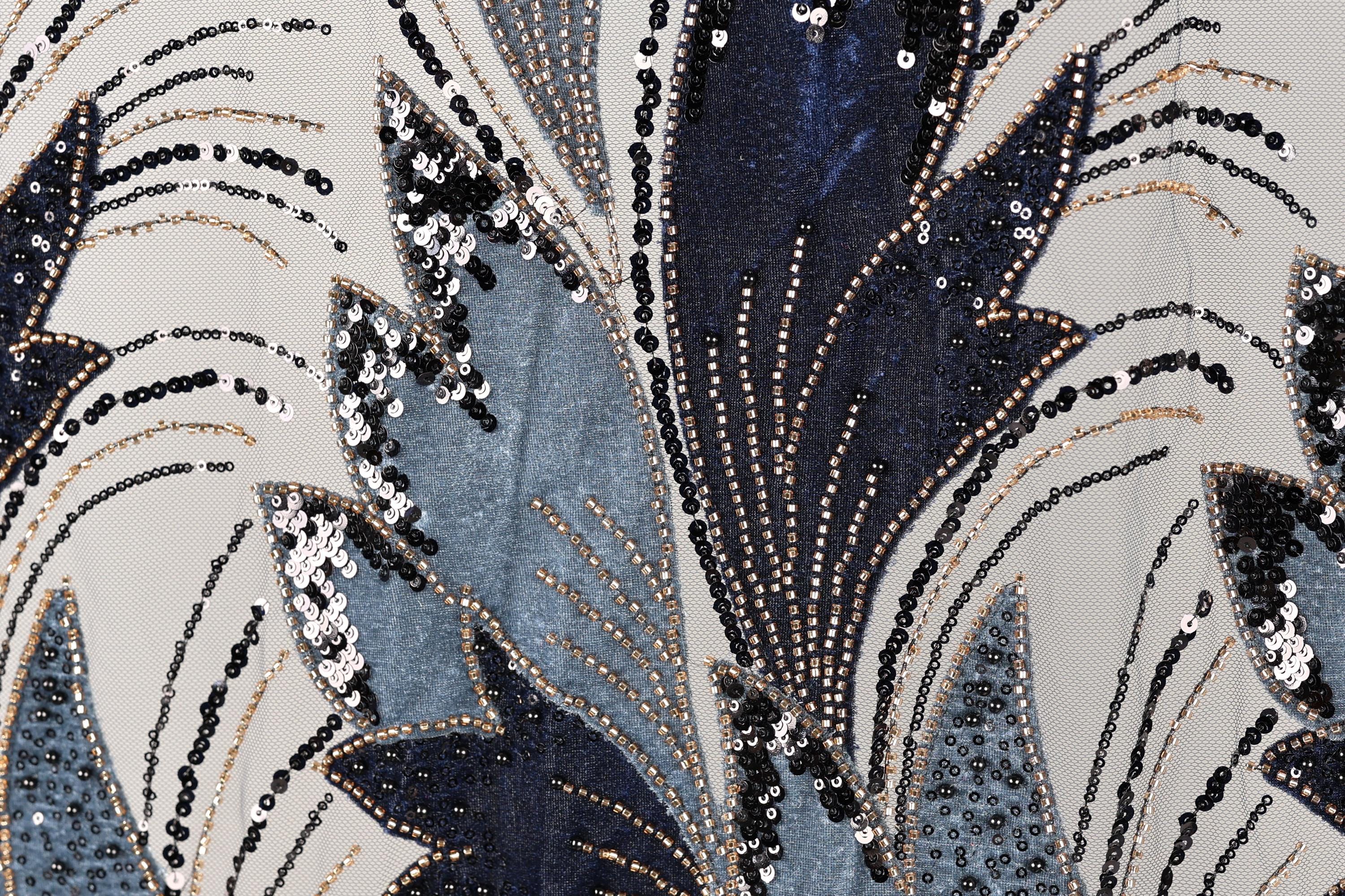 The image is a close-up of a fabric with a striking pattern of sequins and beads in shades of blue, black, and gold. The pattern is intricate and detailed, with the sequins and beads arranged in a way that creates a visually captivating design.