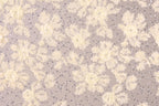 a close-up view of a fabric with a floral pattern, including white and beige flowers and small silver sequins or beads scattered throughout the fabric.