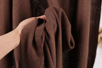 A hand is holding a brown fabric, possibly a piece of clothing or a textile, in front of a brown curtain.