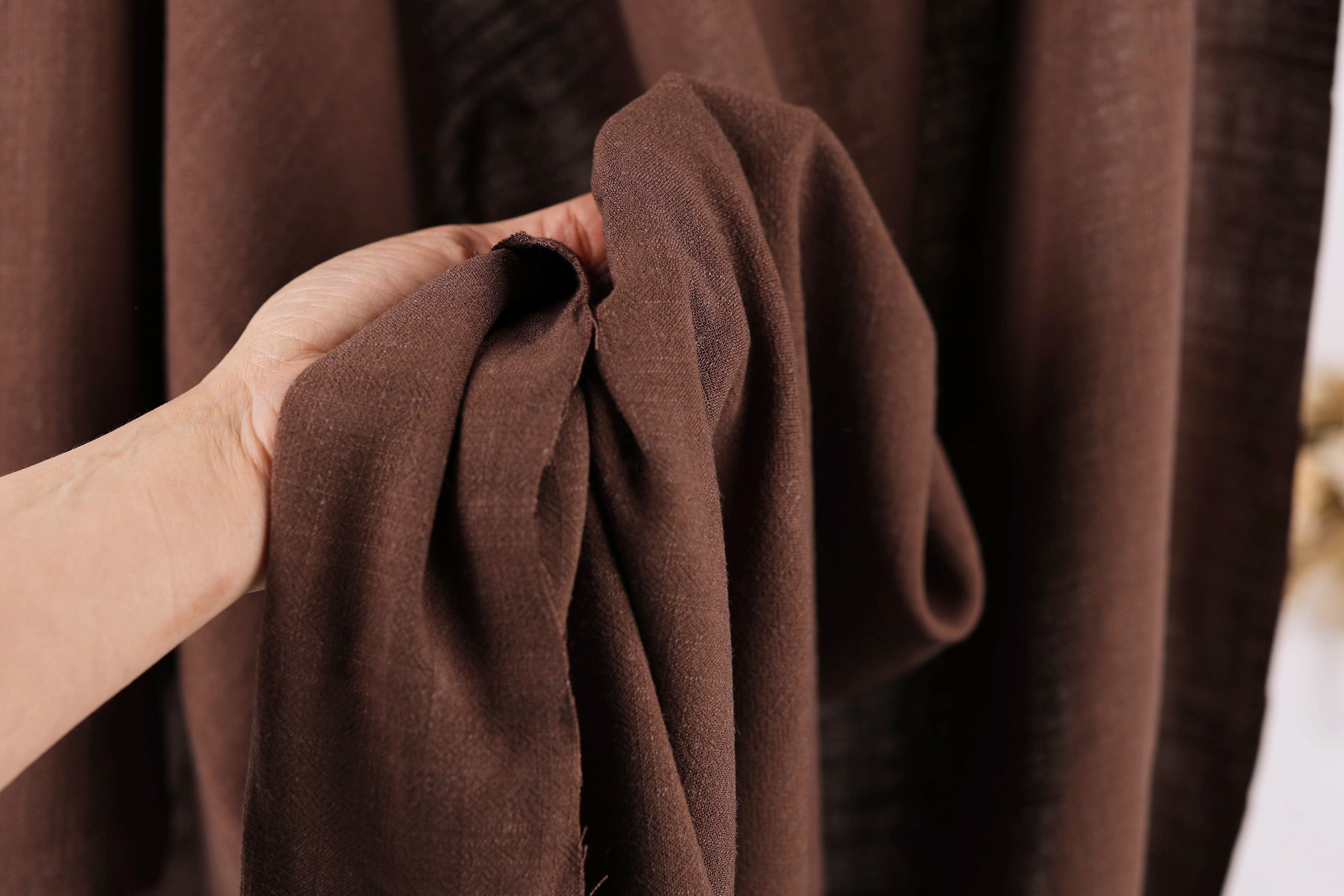 A hand is holding a brown fabric, possibly a piece of clothing or a textile, in front of a brown curtain.