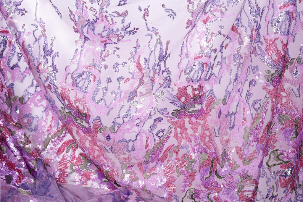 The image is a close-up view of a fabric with a vibrant, multicolored floral pattern. The fabric appears to be made of a sheer material, with a mix of pink, purple, and green colors. The pattern is intricate and detailed, featuring various shapes and textures that create a visually striking and eye-catching design.