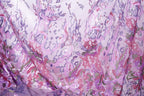 The image is a close-up view of a fabric with a vibrant, multicolored floral pattern. The fabric appears to be made of a sheer material, with a mix of pink, purple, and green colors. The pattern is intricate and detailed, featuring various shapes and textures that create a visually striking and eye-catching design.