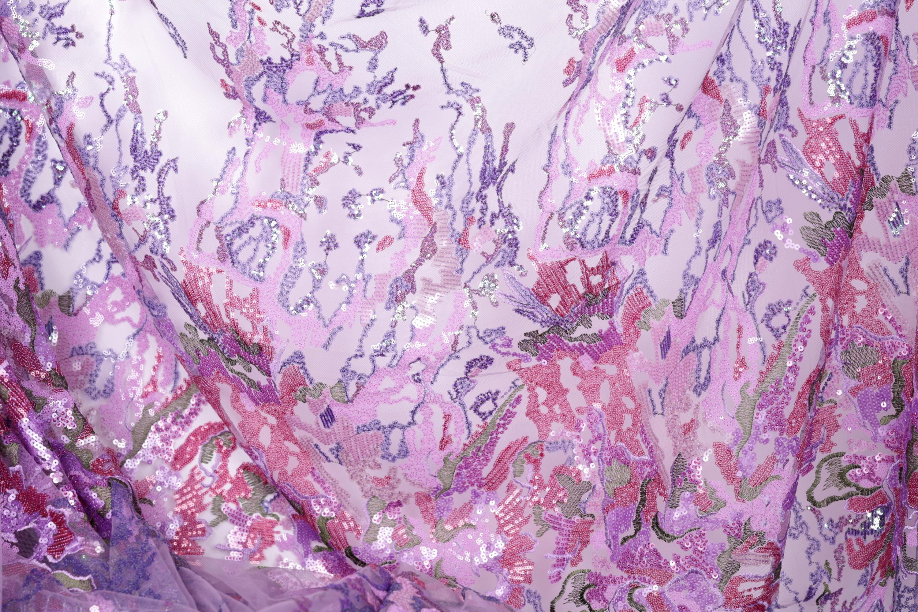 The image is a close-up view of a fabric with a vibrant, multicolored floral pattern. The fabric appears to be made of a sheer material, with a mix of pink, purple, and green colors. The pattern is intricate and detailed, featuring various shapes and textures that create a visually striking and eye-catching design.
