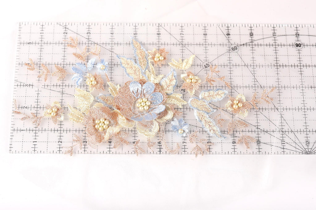 a close-up view of a clear ruler with a floral embroidery design on it. The design consists of various flowers and leaves in shades of blue, pink, and white, as well as some pearls. The ruler serves as a guide for the embroidery, helping to ensure accurate measurements and alignment of the floral elements.