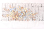 a close-up view of a clear ruler with a floral embroidery design on it. The design consists of various flowers and leaves in shades of blue, pink, and white, as well as some pearls. The ruler serves as a guide for the embroidery, helping to ensure accurate measurements and alignment of the floral elements.