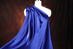 A mannequin wearing a flowing, one-shoulder blue satin garment against a dark background.