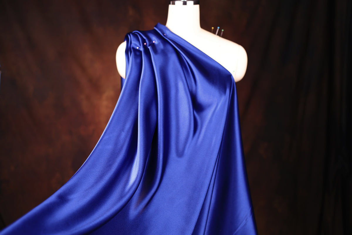 A mannequin wearing a flowing, one-shoulder blue satin garment against a dark background.
