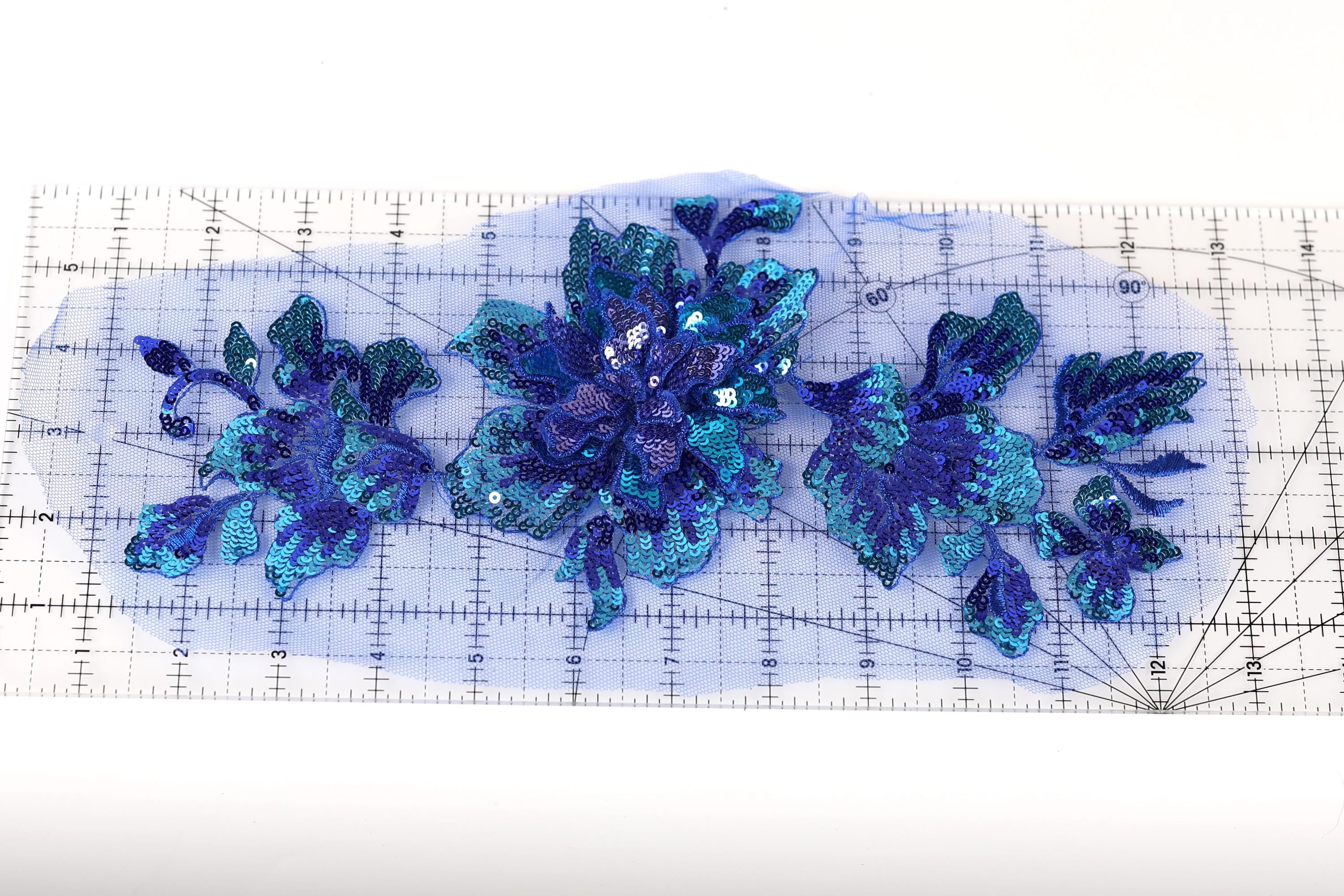 a close-up view of a piece of fabric with a blue and green sequined flower design. The fabric appears to be a light blue color, and the flower design is composed of small sequins in shades of blue and green. The fabric is laid out on a transparent surface, which allows for a clear view of the intricate details of the sequined design.
