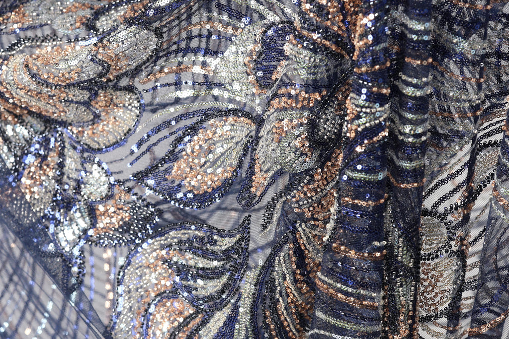 The image is a close-up of a fabric with a complex, intricate pattern of sequins and beads in shades of blue, gold, and silver. The pattern appears to be a floral or leaf-like design, with the sequins and beads arranged in a way that creates a visually striking and detailed appearance.